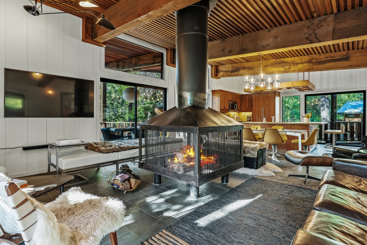 A central open hearth fireplace draws attention, surrounded by an open living space that features wooden beams and large windows. A cozy seating area includes a stylish lounge chair and a sofa, while a modern kitchen is visible in the background.