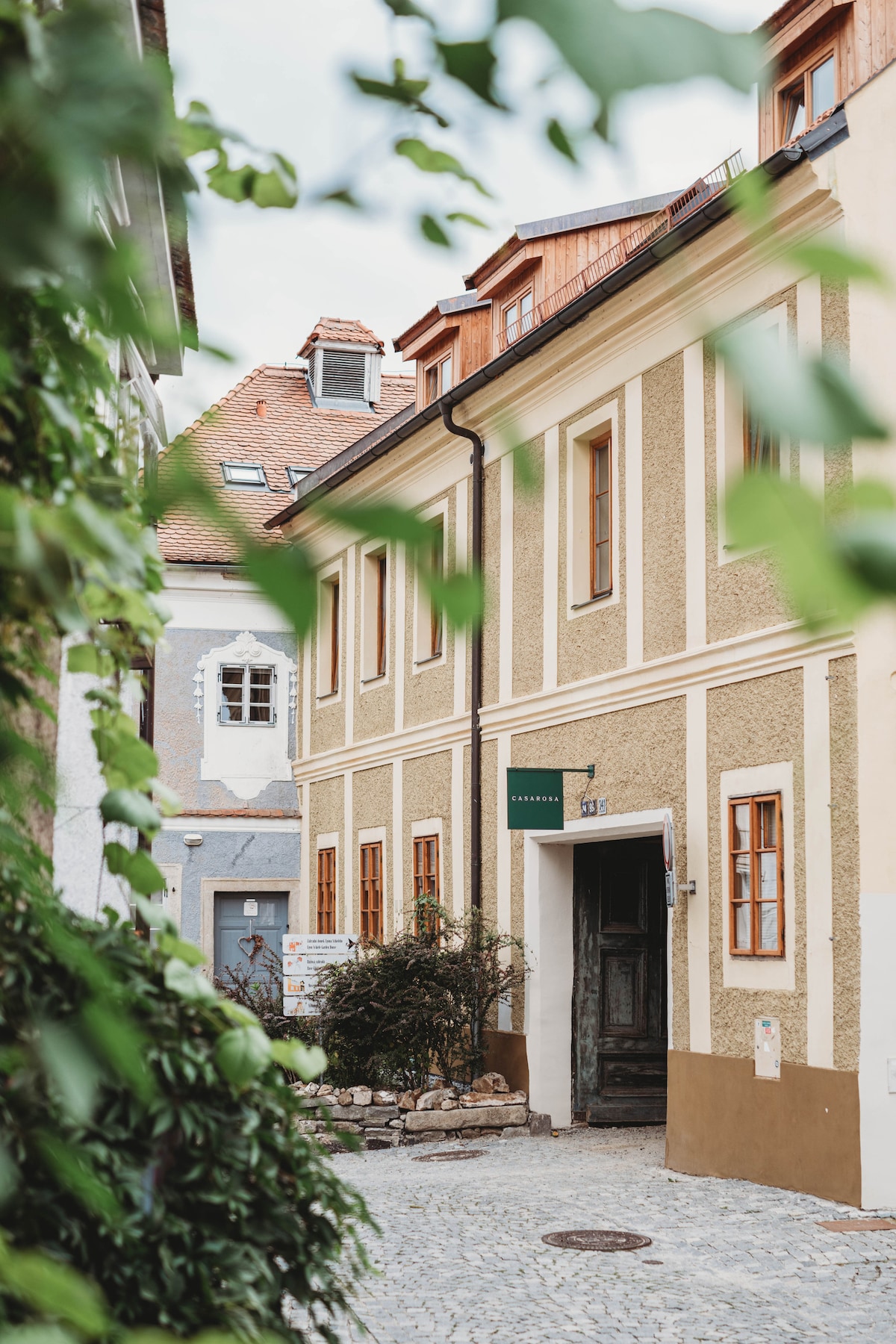 A charming building exterior is shown, featuring textured yellow walls and red roof accents. Green foliage frames the scene, highlighting the inviting entrance with wooden doors. Cobblestone pathways are visible, contributing to the historic ambiance of the surrounding area.