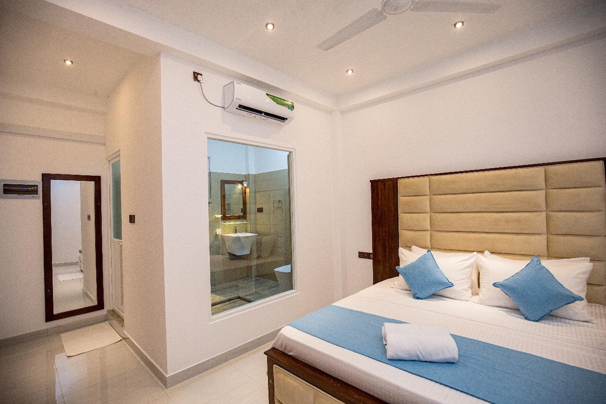A well-lit room features a spacious bed adorned with light blue decorative pillows. A glass-enclosed bathroom is visible, showcasing modern fixtures. Soft lighting enhances the clean, white walls, and a ceiling fan contributes to the room's air circulation.