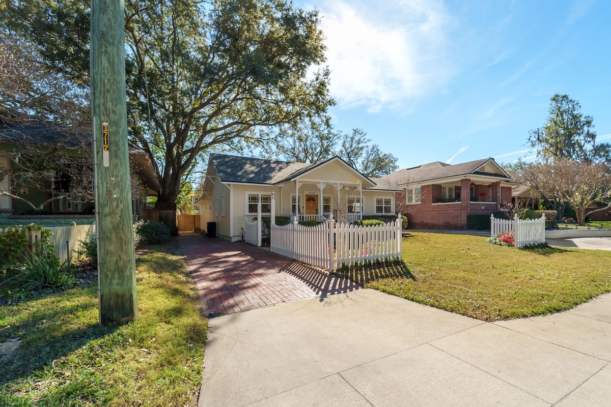 A one-story home features a welcoming front porch surrounded by a white picket fence. The driveway provides parking space for vehicles, leading to a well-maintained yard with lush greenery. Large windows allow natural light into the living area, showcasing a bright and open ambiance.