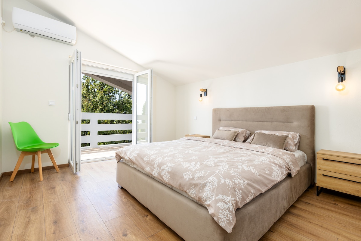 A spacious sleeping area features a comfortable bed with soft bedding and two nightstands beside it. Large windows open to a private terrace, allowing natural light to brighten the room. A modern chair in the corner adds a touch of color to the wood floor.