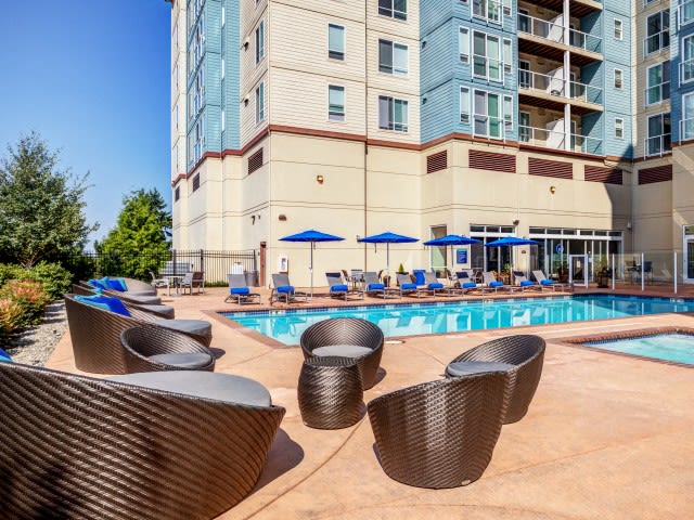 An outdoor pool area is depicted, surrounded by modern lounge chairs in a woven texture. Bright blue umbrellas provide shade, enhancing the inviting atmosphere. The pool reflects sunlight, and the backdrop features an apartment building with multiple levels, contributing to a spacious recreational environment.
