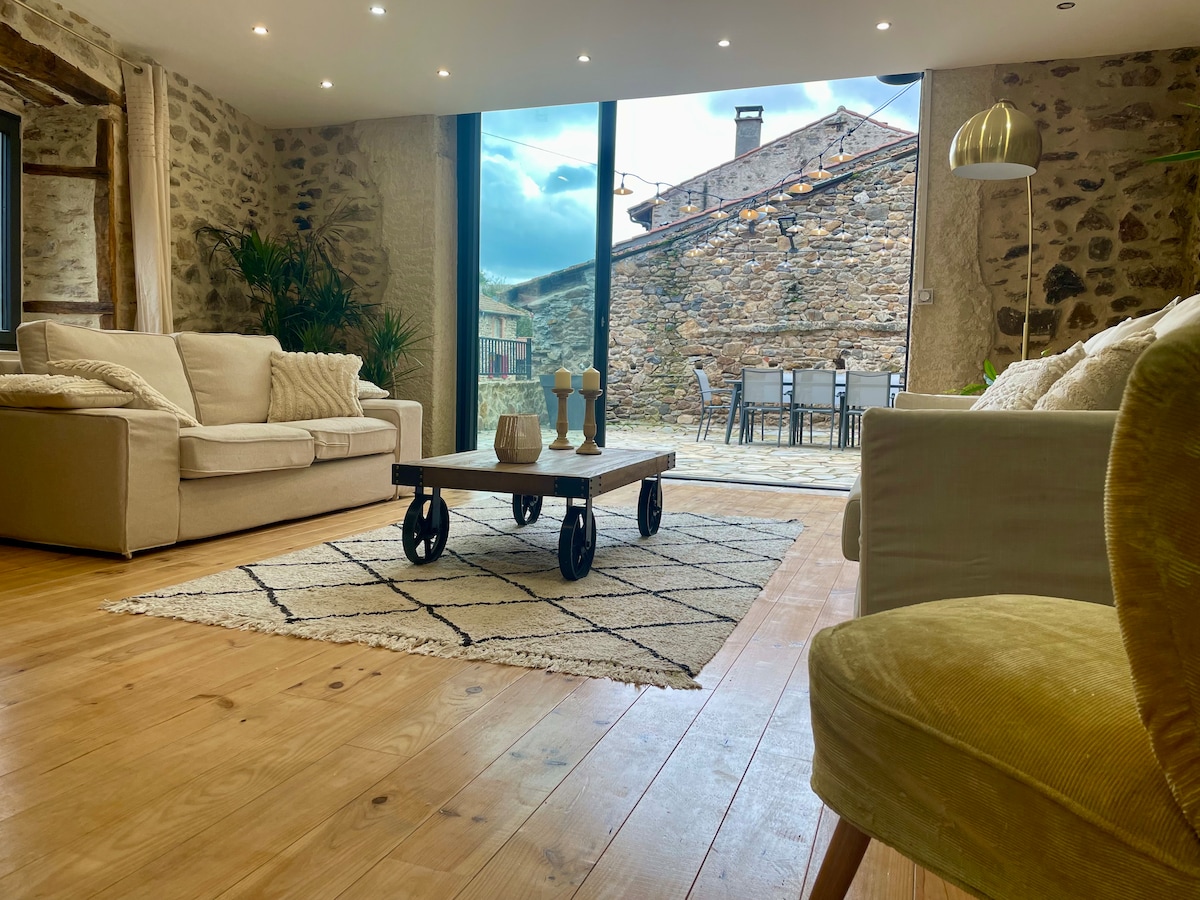 A spacious living area features two soft sofas, including a light-colored one and a green armchair. A coffee table stands at the center on a patterned rug, with two decorative candles displayed on top. Large glass doors open to an outdoor terrace, allowing natural light to fill the room.