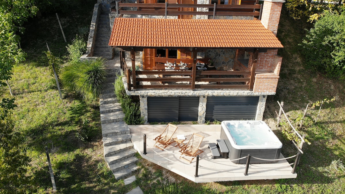 An aerial view of the villa reveals a spacious outdoor terrace with a hot tub and two wooden lounge chairs. Steps lead down to a well-maintained garden area, showcasing greenery and a rustic charm. The villa's wooden deck offers ample space for dining and relaxation.