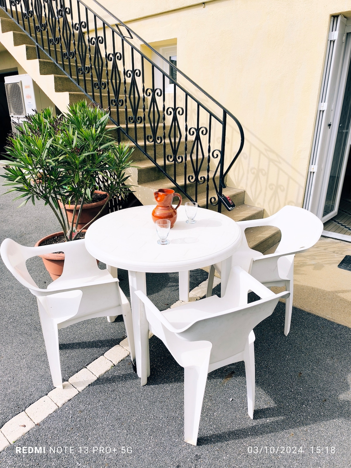 An outdoor seating area is featured, consisting of a round white table accompanied by four white plastic chairs. A terracotta jug and two glasses sit on the table. Lush green plants are placed nearby, with a staircase in the background leading to the entrance.