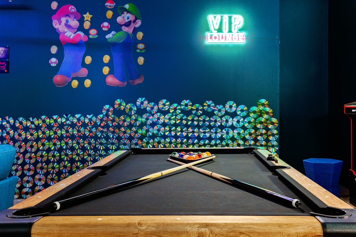 A vibrant game room is featured, with a billiard table in the foreground and retro-themed wall art depicting iconic video game characters. A neon 'VIP Lounge' sign glows against a dark wall, complemented by colorful circular decor made of CDs, creating a playful atmosphere.