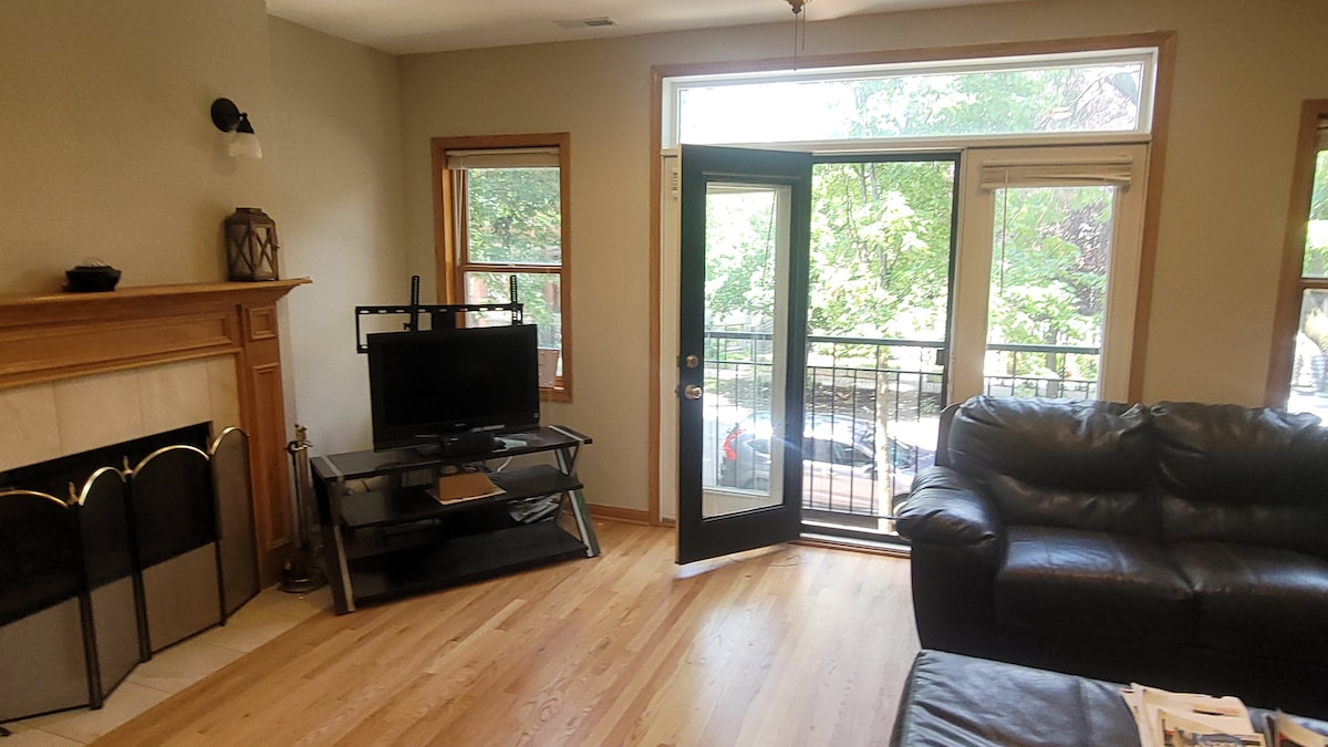 A living area features a black leather sofa and a glass TV stand, with a television in view. Natural light enters through windows, and a balcony door opens to outdoor greenery. The wooden flooring adds warmth to the space.