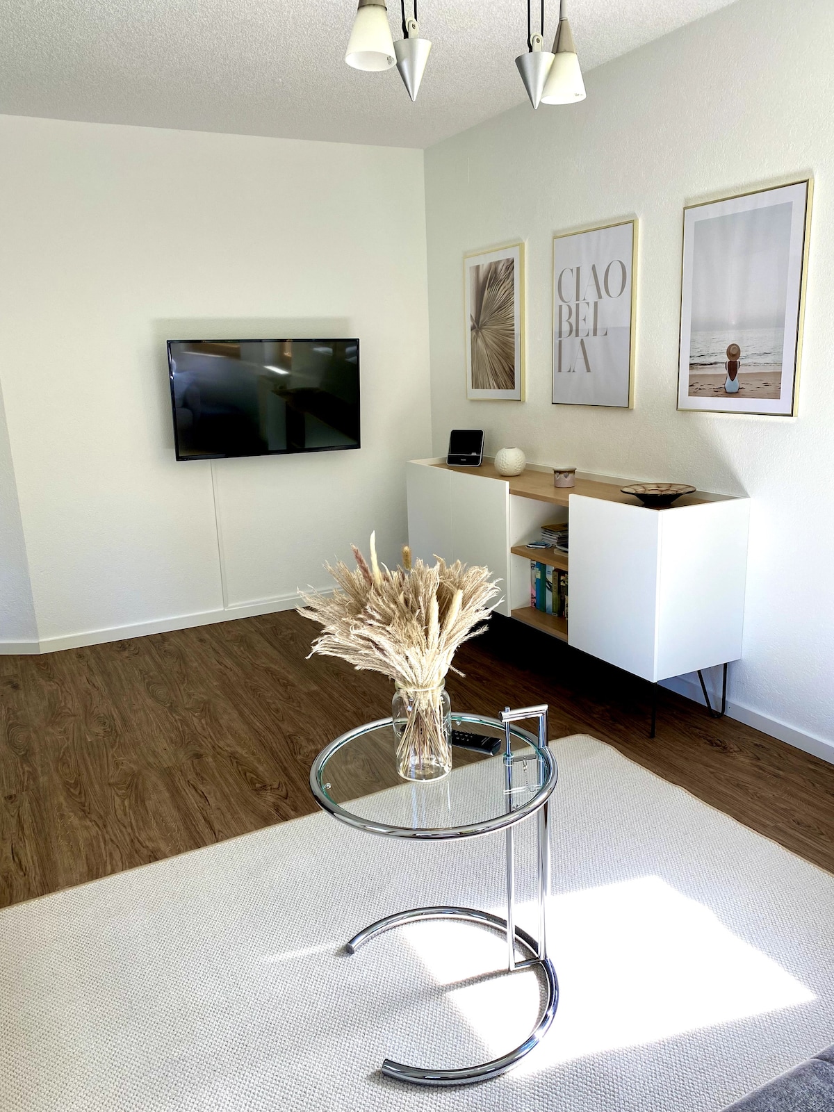 A living area is presented with a mounted flat-screen TV and light-colored walls. A white sideboard displays decorative items and books. A round glass side table is positioned beside a textured area rug, while artwork in neutral tones adorns the walls.