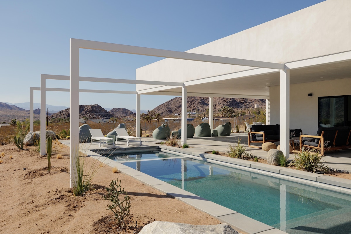 A modern home features a sleek pool surrounded by minimal landscaping, with lounge seating and shaded outdoor living areas visible. The desert landscape showcases rocky hills and dry vegetation, extending into the distance under a clear blue sky.