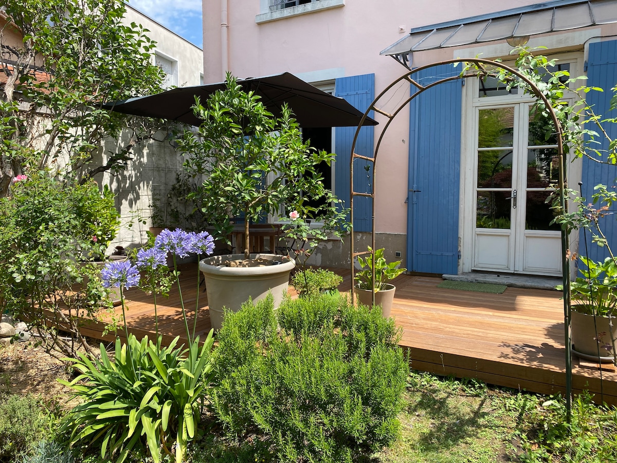 A private terrace is showcased, featuring a wooden deck surrounded by greenery. A small fountain is visible in the center, complemented by flowers and potted plants. Blue shutters frame the entry to the apartment, enhancing the tranquil atmosphere of the outdoor space.