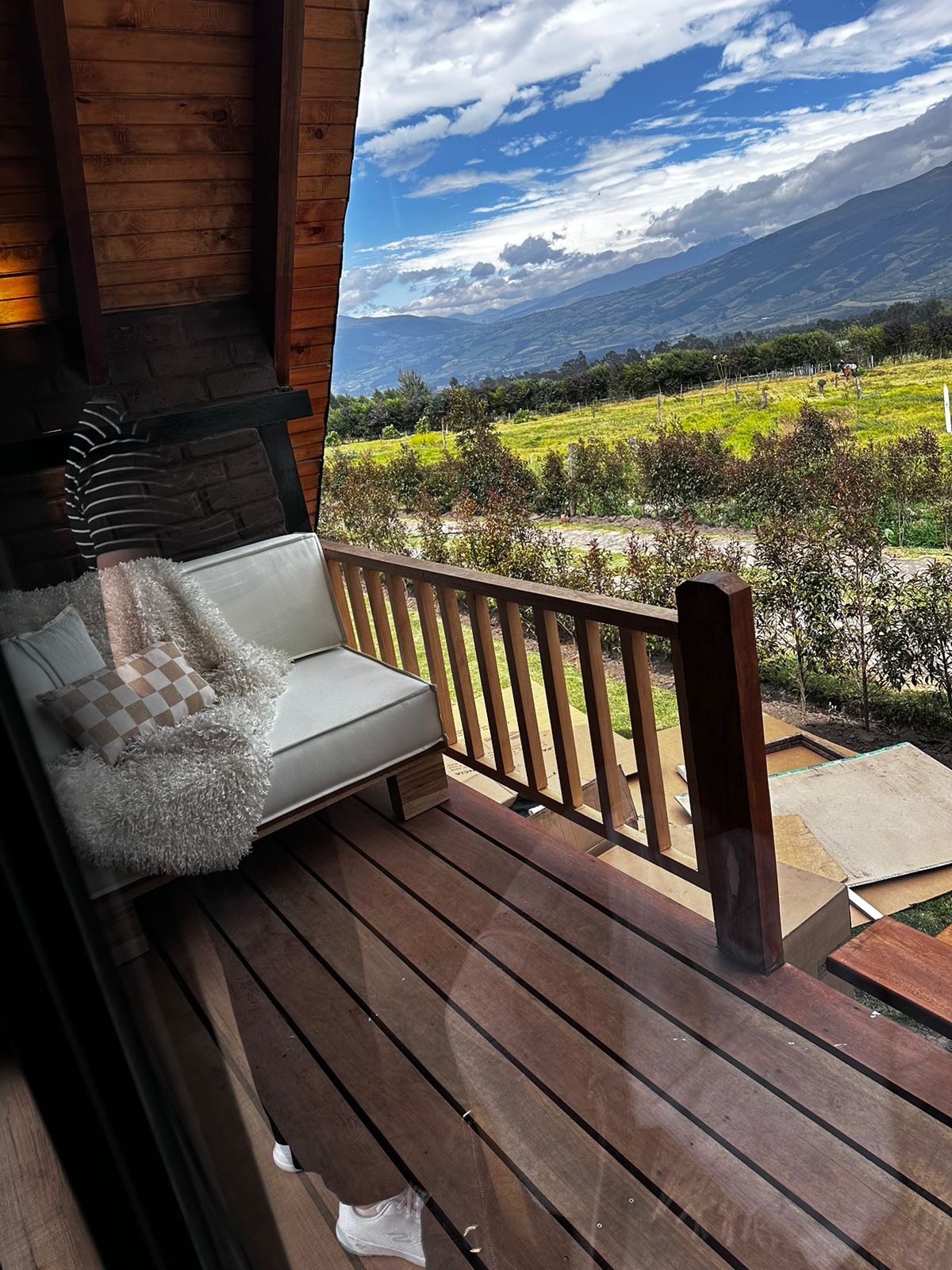 A furnished balcony features a white couch adorned with cushions, surrounded by wooden railing. Lush greenery and a sweeping landscape are visible in the background under a bright blue sky, providing an inviting space for relaxation.