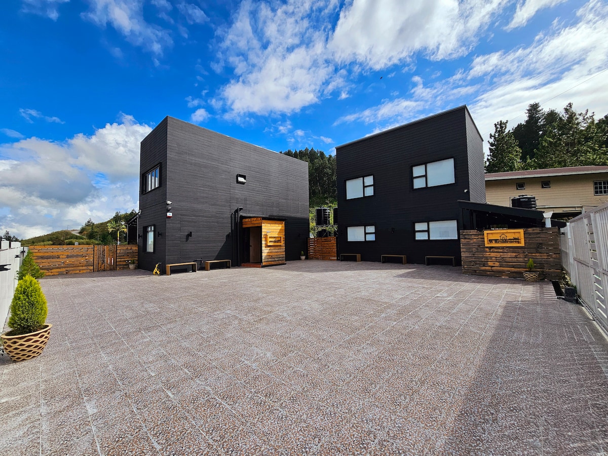 An exterior view of the property showcases a contemporary design with two distinct buildings. A spacious parking area is visible, surrounded by a clean stone surface. Clear blue skies and scattered clouds provide a serene backdrop.