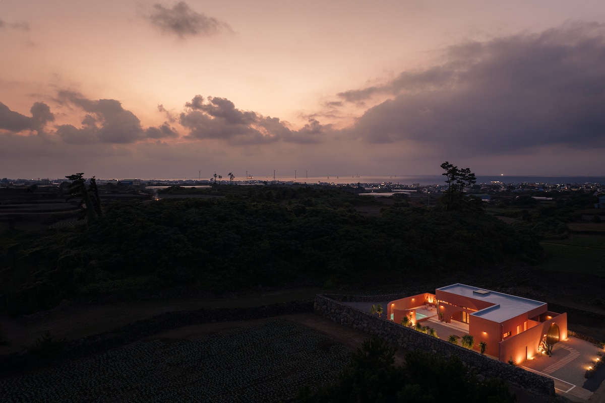 An evening view captures the villa situated amidst natural greenery, illuminated by soft lights. The horizon blends with a dusky sky, creating a serene atmosphere. The distant coastline and gentle hills are visible, enhancing the villa's sense of seclusion and tranquility.