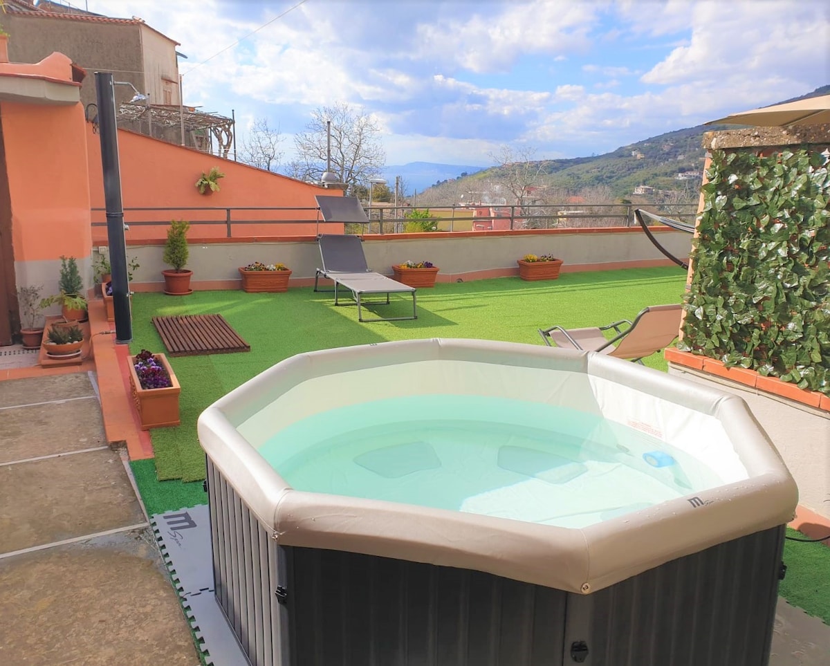 A spacious private terrace features a circular hot tub surrounded by greenery and comfortable sun loungers. Potted plants line the terrace, and the area is bathed in natural light, with distant hills visible under a partly cloudy sky.