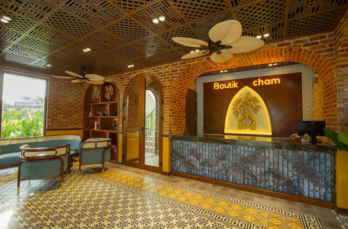 The reception area features a combination of brick walls and intricate tile flooring, complemented by wooden accents. A curved counter with a glowing sign displays the name 'Boutik Cham.' Two ceiling fans circulate air in the open space, which is filled with natural light from large windows.