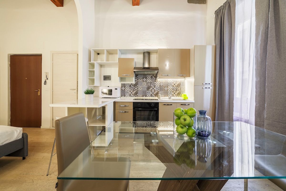 A well-equipped kitchen and dining area are visible, featuring modern appliances and a decorative backsplash. A glass dining table is set, with a bowl of green apples adding a touch of color. Soft natural light filters through sheer curtains, enhancing the open and airy feel.