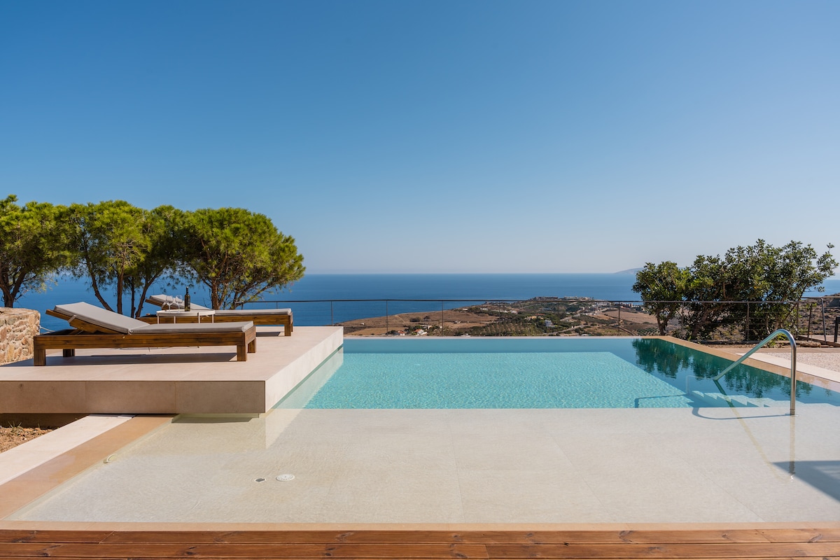 A private swimming pool is shown with a modern edge, overlooking a sea view. Sun beds are positioned to the side on a wooden deck, surrounded by lush green trees. The clear blue sky complements the tranquil atmosphere of the outdoor area.