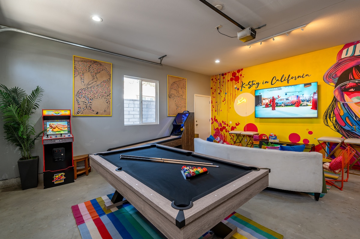 A vibrant game room is presented, featuring a large pool table at its center. An arcade game machine stands to one side, while a bright yellow wall displays colorful artwork. A comfortable sectional sofa is arranged for relaxation, and a TV is mounted on the wall.
