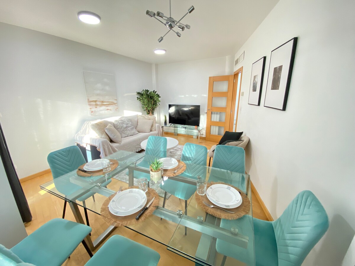 A spacious living area features a glass dining table set for six, accompanied by stylish turquoise chairs. A comfortable sofa is positioned nearby, facing a wide-screen TV. Neutral wall art and a decorative plant add a touch of elegance to the light-filled space.