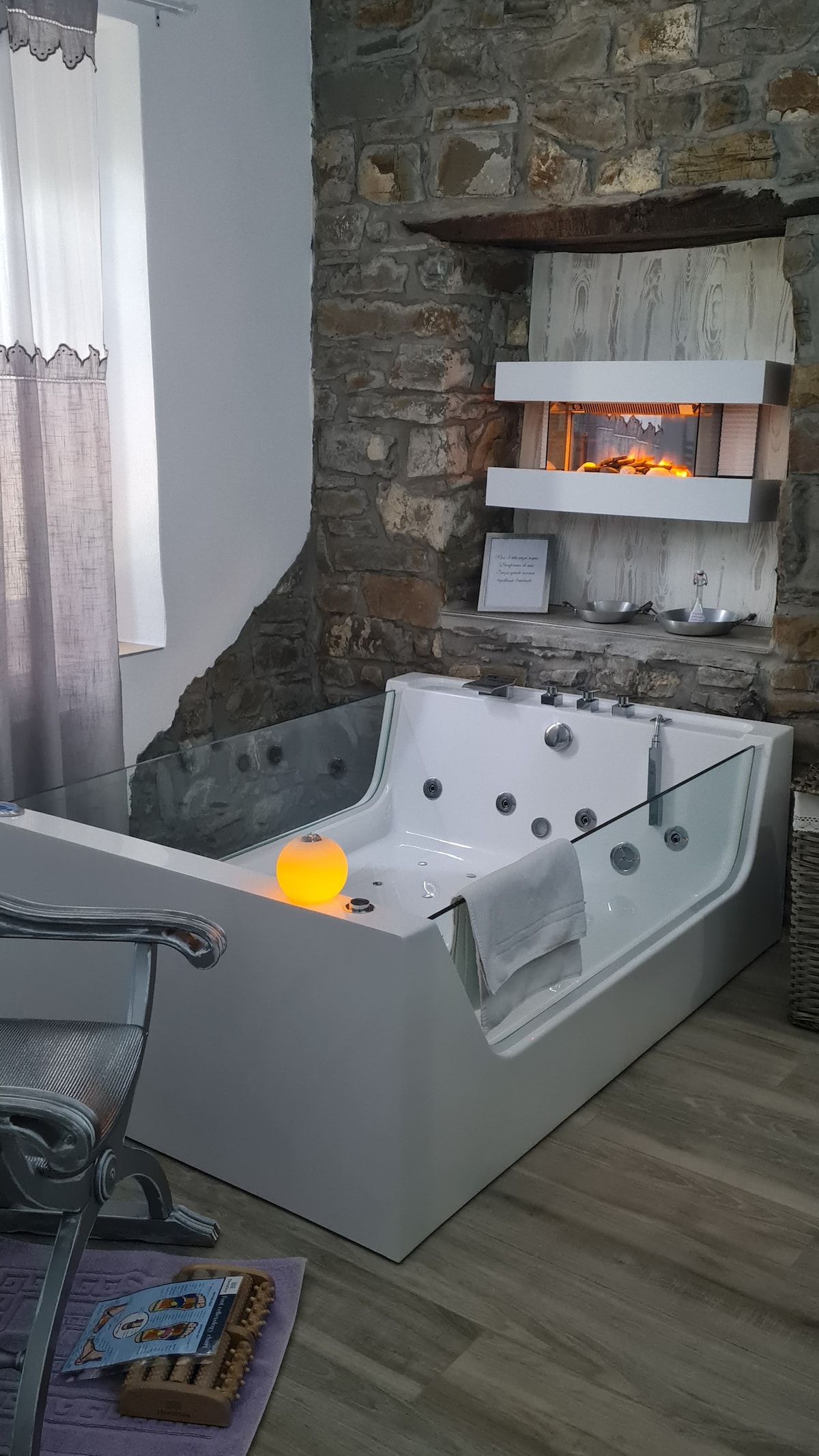 A wellness bathroom features a spacious jetted tub with a glass panel for natural light. A subtle fireplace is positioned above, complemented by stone walls. A soft glow is emitted from an orb lamp nearby, while a plush seat and decorative items add to the serene setting.