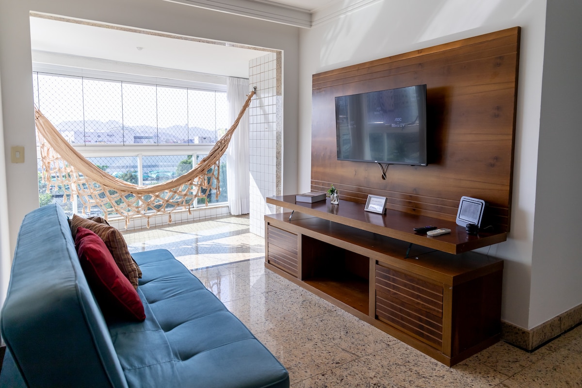 A well-lit living area showcases a comfortable blue sofa accented with red cushions. A modern entertainment unit is positioned against the wall, featuring a flat-screen television. Natural light filters through large windows, illuminating a balcony with a hammock.