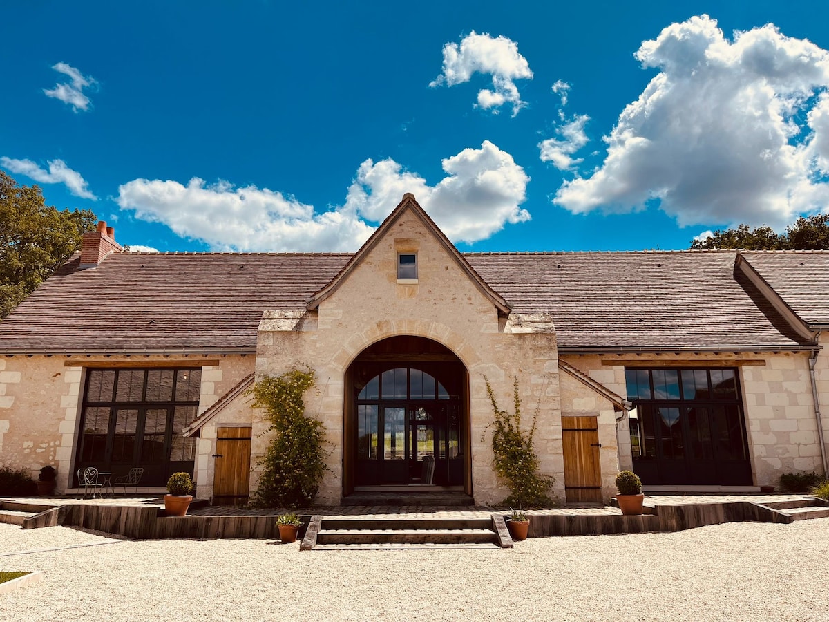 The charming exterior of the house showcases a blend of traditional stone and modern design elements. Large glass doors are framed by wooden accents, enhancing the inviting entrance. Lush greenery and decorative pots line the walkway, set against a clear blue sky adorned with clouds.