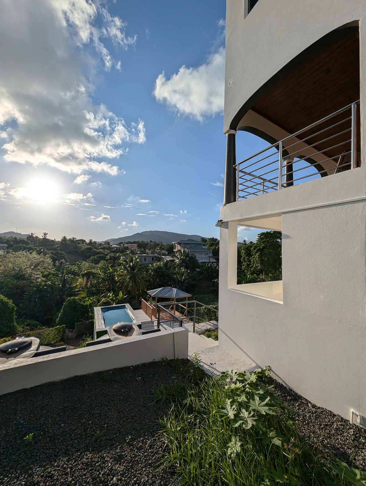 The exterior of the building features a modern aesthetic with arched windows and a view of the surrounding greenery. A deck area and a small pool are visible in the background, while the sun casts a warm light over the scene, highlighting the natural landscape.