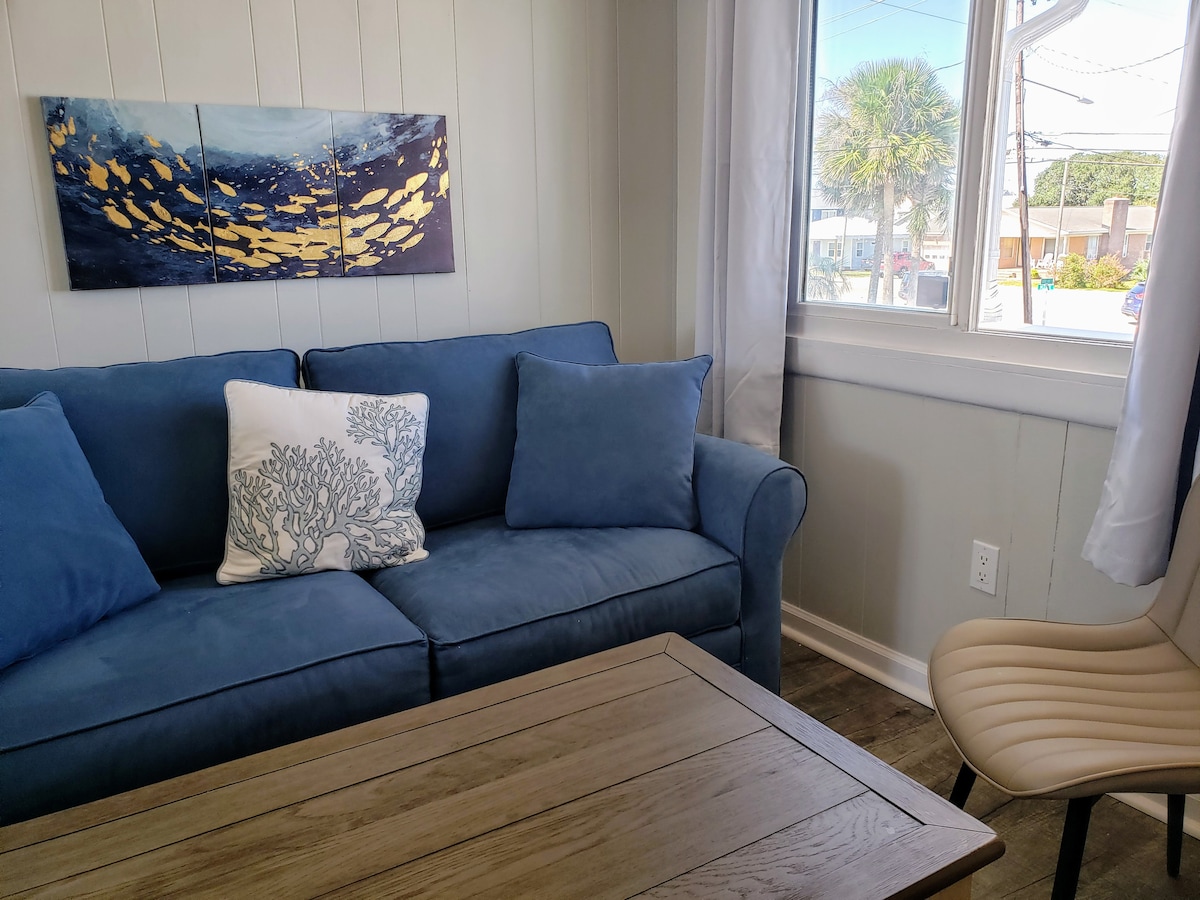 A comfortable love seat in a coastal-themed room is paired with a lift-top coffee table. Natural light filters through large windows, illuminating the space. A wall-mounted artwork featuring golden fish adds a maritime touch, enhancing the beach town vibe.