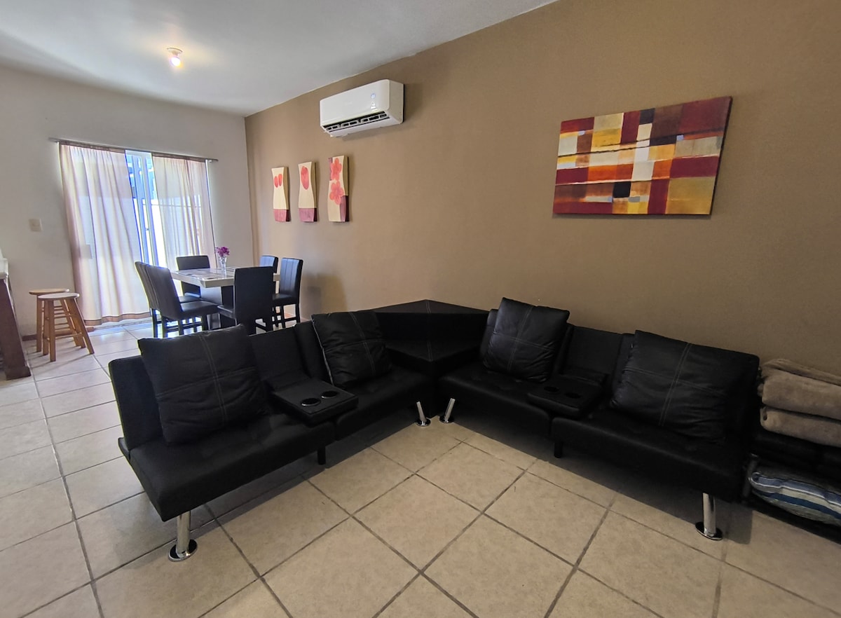 The living area features a modern black sectional sofa with built-in cup holders, positioned against a warm-toned wall. Two pieces of abstract wall art provide color, while a dining table with chairs is visible in the background, next to a set of sliding glass doors allowing natural light.