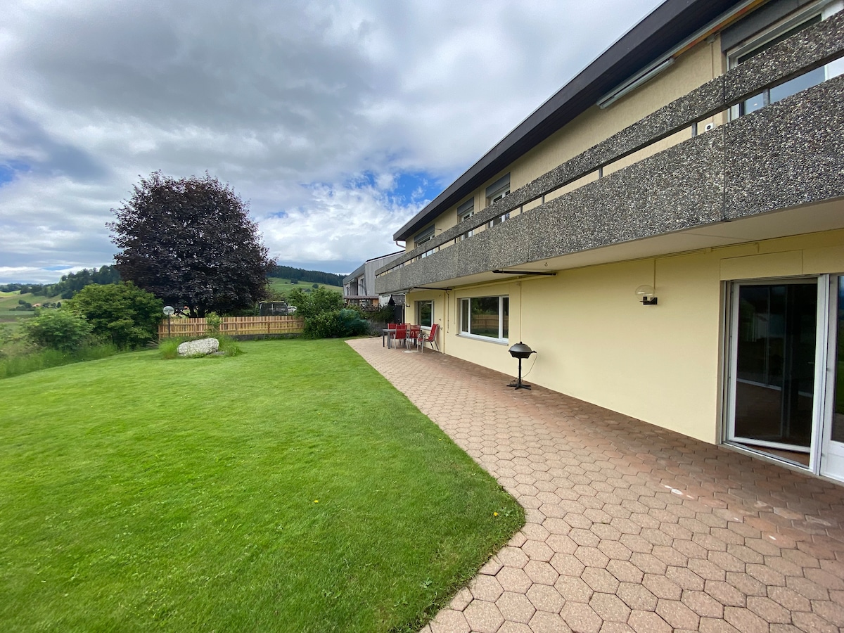 A spacious outdoor patio area is shown, featuring a well-maintained lawn with a barbecue grill. Tables and chairs are positioned nearby, set against a backdrop of greenery and distant hills. The sky is partly cloudy, creating a serene outdoor environment.
