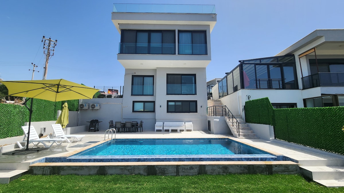 The two-story villa features a private pool surrounded by a spacious sun terrace. Lounge chairs are arranged near the pool, complemented by an umbrella for shade. A green hedge provides privacy, while the modern glass facade of the villa reflects the bright blue sky.