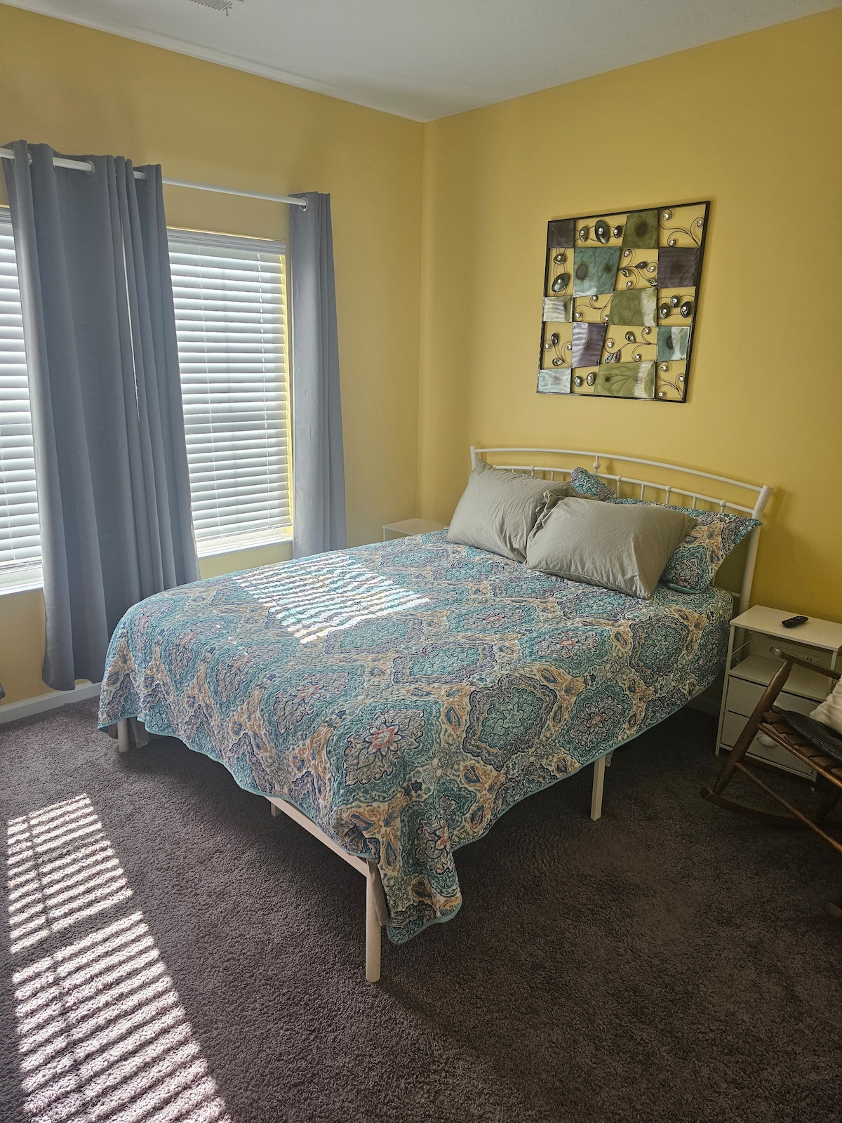 A cozy bedroom is highlighted by yellow walls and natural light streaming through window blinds. A spacious bed is covered with a decorative blue and beige bedspread. Two gray curtains frame the window, and framed wall art adds a personal touch to the inviting space.