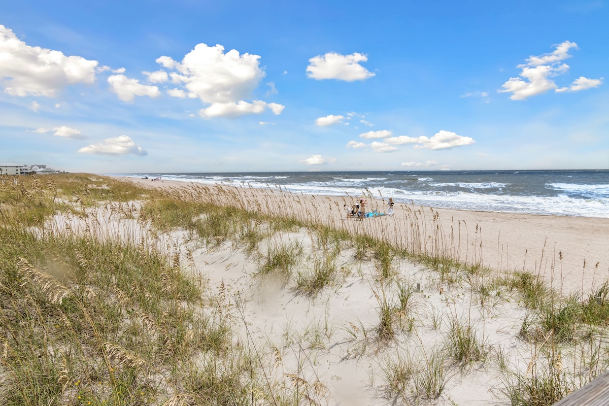 A stretch of sandy beach is bordered by tall grasses gently swaying in the breeze. The Atlantic Ocean meets the shore with rolling waves under a bright blue sky, dotted with fluffy white clouds. Sunbathers and beachgoers can be seen in the distance enjoying the serene setting.