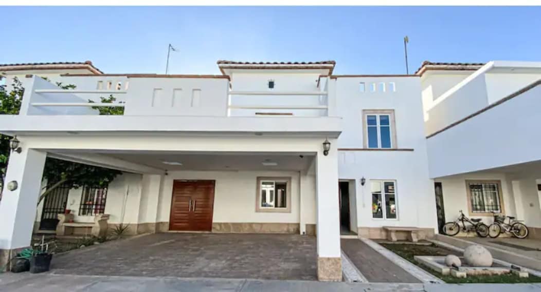 A modern residential building features a spacious front facade with a wooden door as its focal point. Large windows are present throughout, enhancing natural light. The area includes a driveway and paved pathways, alongside greenery that adds a touch of nature to the setting.