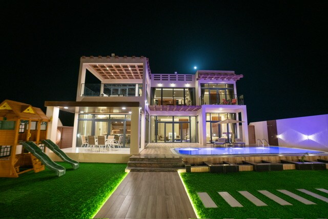 The modern villa is showcased at night, featuring multiple levels and large glass windows. A spacious terrace includes a swimming pool and lounge areas. Illuminated landscaping frames the structure, while a playset and slides are visible in the yard, enhancing the outdoor space.
