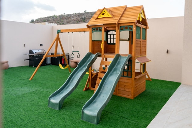 An outdoor play area features a wooden playhouse with a slide and swings. A grassy surface surrounds the equipment, creating a family-friendly space. A barbecue grill is positioned nearby, providing an option for outdoor cooking.