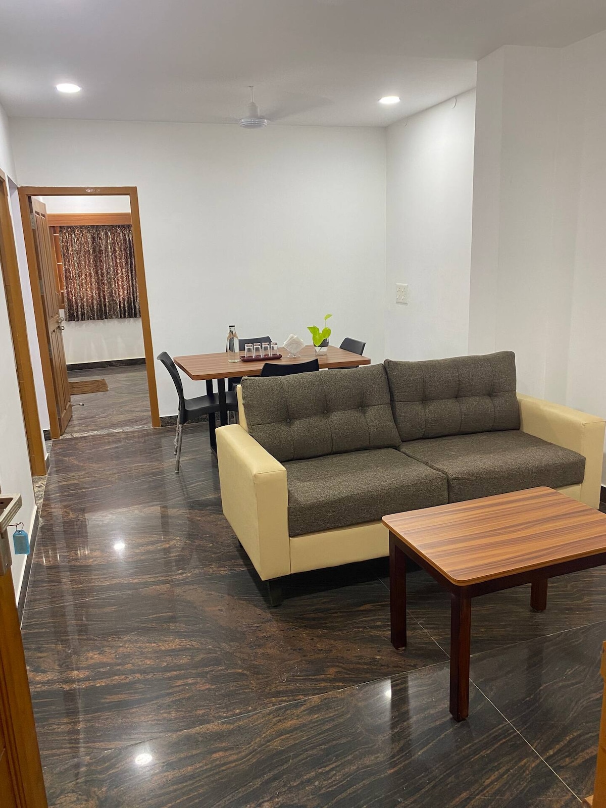 A modern living area features a neutral-colored sofa with a dark cushion. A wooden coffee table sits in front of the seating area. In the background, a dining table with chairs is visible, along with a doorway leading to another room.