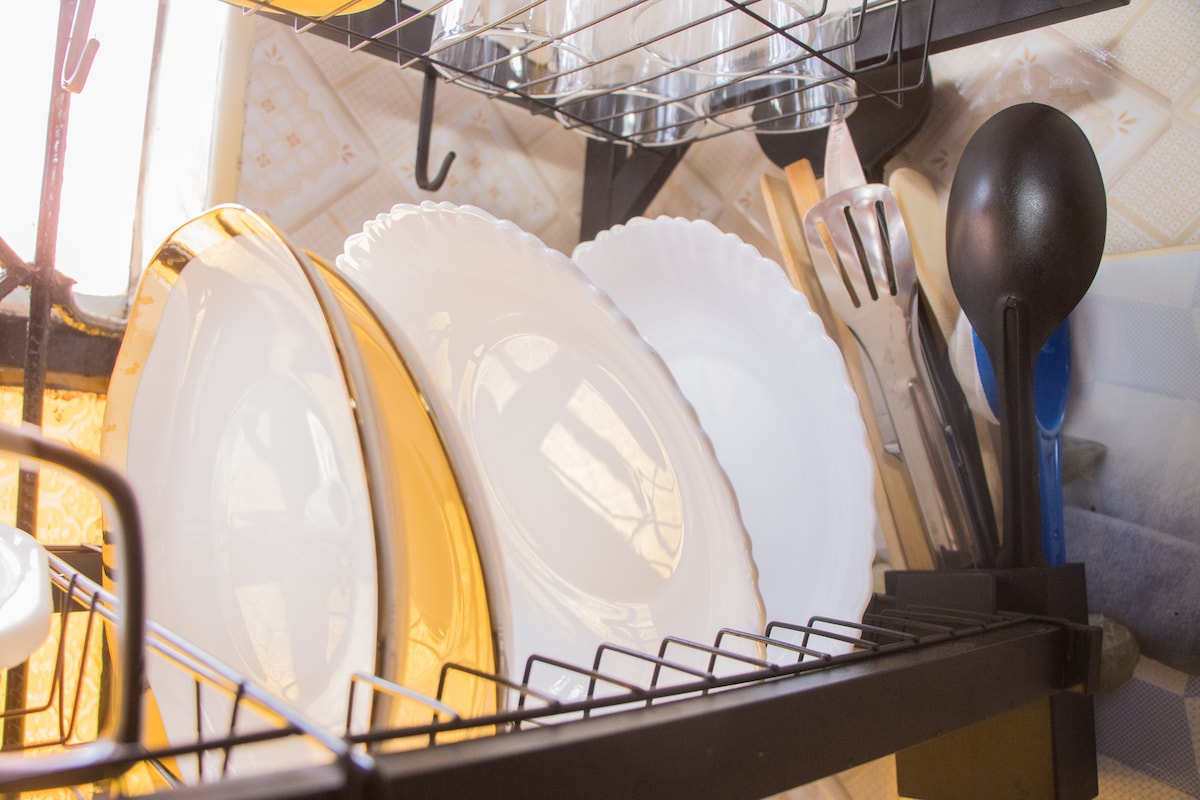 A metal shelf holds an assortment of dishes and utensils. The arrangement includes white plates and shiny gold-rimmed plates, along with various kitchen tools and glassware, creating a practical and organized dining setup.