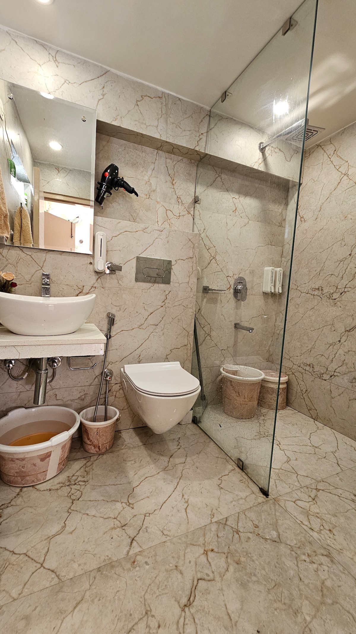 A modern bathroom features marble wall tiles and a spacious glass shower enclosure. A clean, white toilet and a stylish sink are complemented by minimalist decor. Two woven baskets are positioned beside the sink, adding a functional touch to the elegant space.