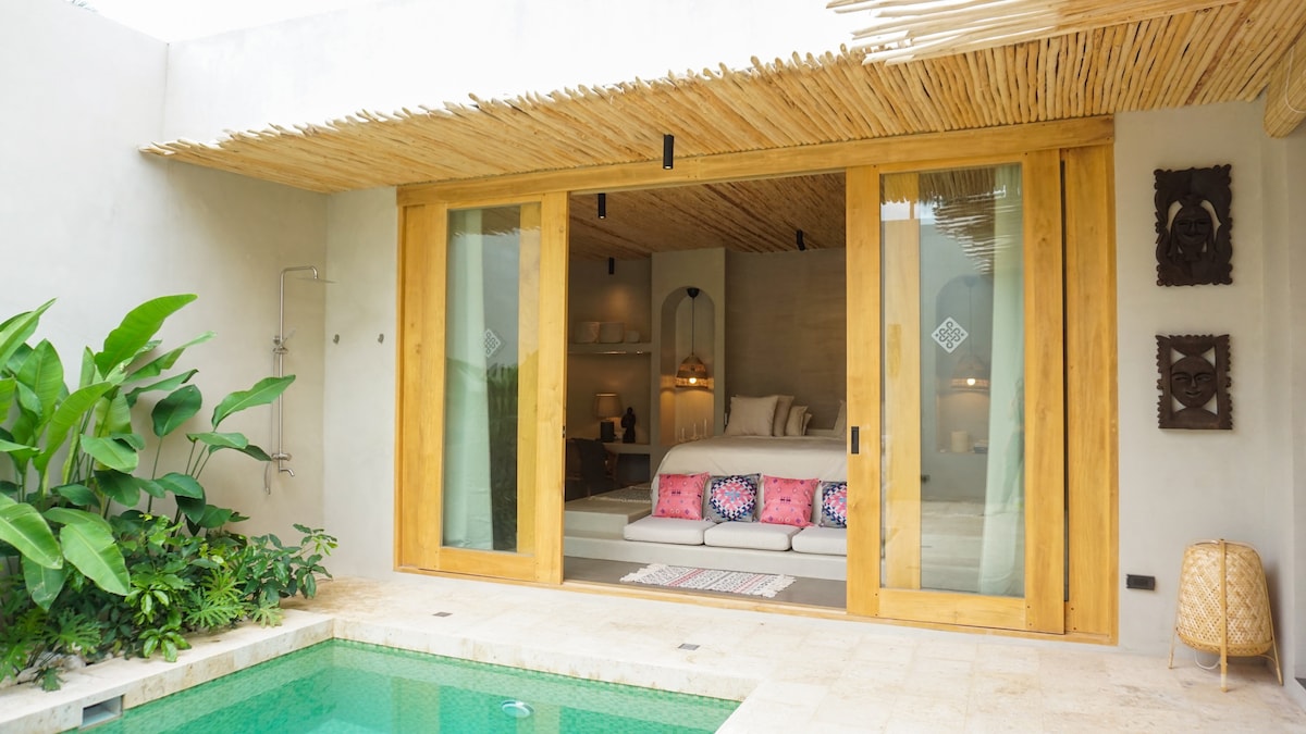 A spacious villa entrance features large glass sliding doors that open to a serene sleeping area. A private plunge pool is visible in the foreground, surrounded by tropical greenery. The bedding is elegantly arranged, and colorful cushions add a touch of vibrancy.