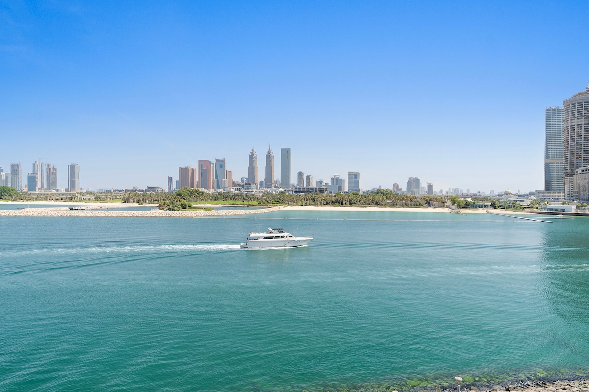 A panoramic view showcases the tranquil waters of the sea with a yacht cruising by. The skyline of Dubai is visible in the background, featuring a mix of modern skyscrapers and palm trees, creating a serene coastal landscape.