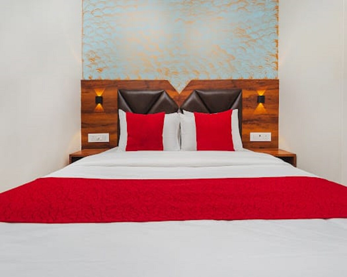 A spacious bed is dressed in crisp white linens with a bold red throw blanket laid across the foot. Two decorative red pillows complement the neutral headboard and wall. Soft lighting is provided by wall sconces on either side, enhancing the overall design.