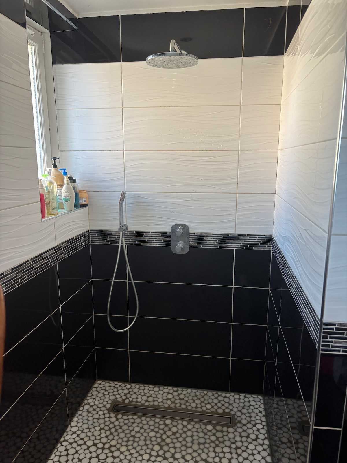 A modern shower space is presented with a sleek design, featuring textured white and black tiles. The showerhead is mounted above, and a handheld option is available. A collection of toiletries is arranged on a small ledge, and a pebble-stone floor adds a natural element.