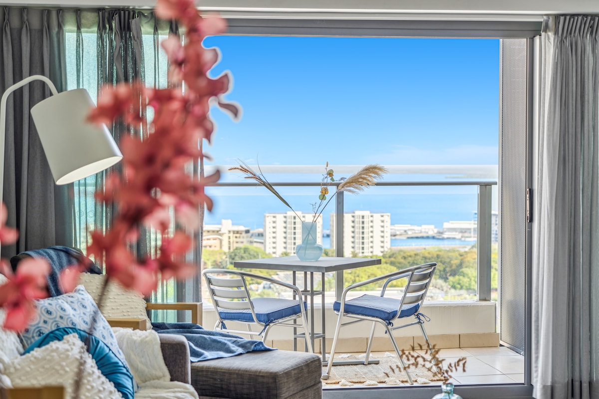 A bright living space features a comfortable sofa adorned with cushions, leading to a breezy balcony. Clear views of the ocean and surrounding buildings are visible through expansive glass doors. A small table is set for two, enhanced by decorative vases and plants.