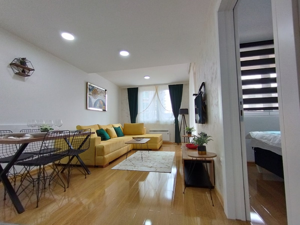 A modern living area showcases a bright yellow sofa complemented by green accent pillows, paired with a glass coffee table. A dining table with metal chairs is positioned nearby, and sunlight filters through sheer curtains, illuminating the room. A bedroom is visible in the background.