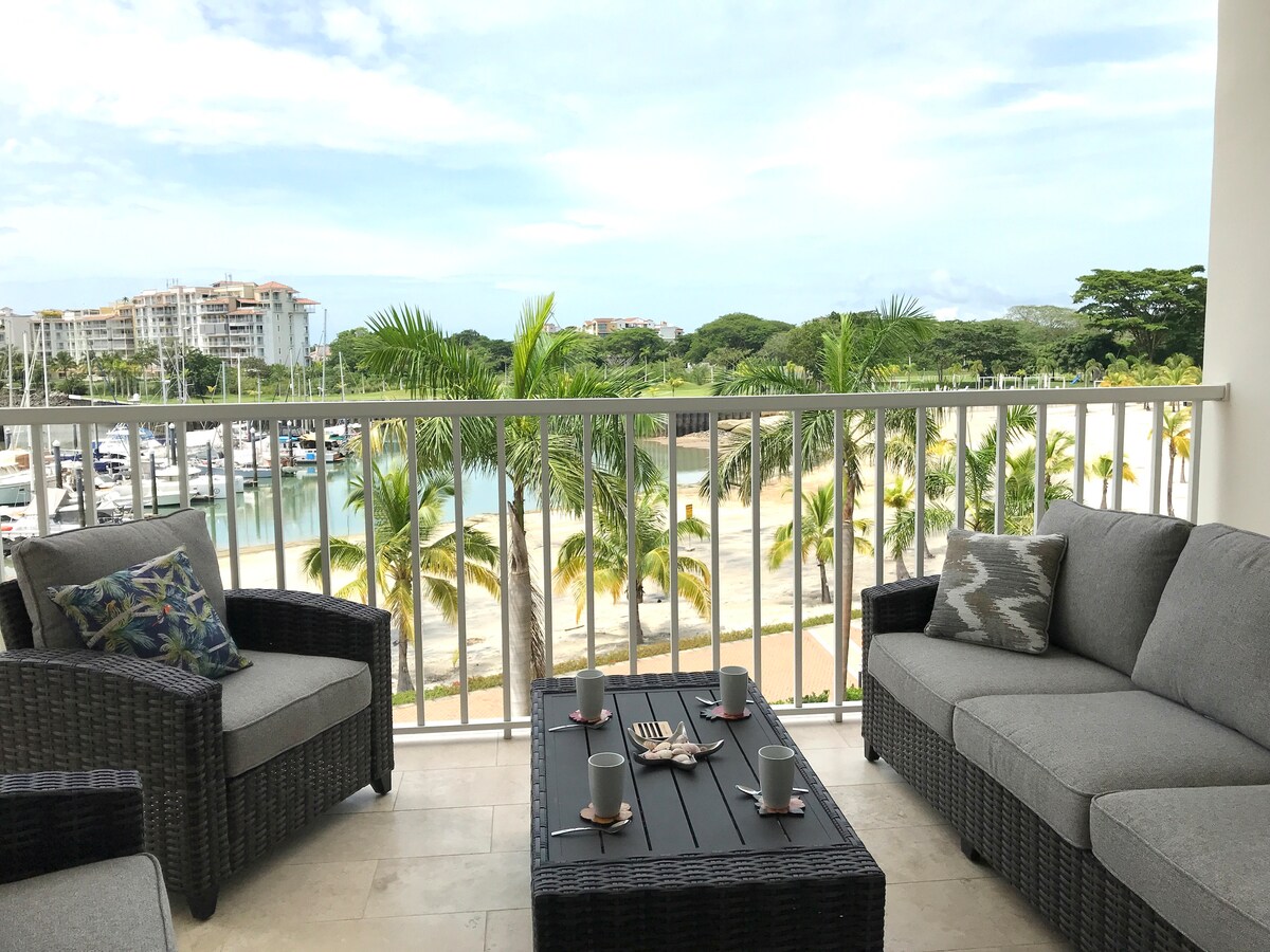A comfortable balcony area features gray woven seating with decorative cushions, positioned around a dark wood table. A scenic marina view is visible beyond the railing, framed by lush palm trees and soft sandy beach. Soft light illuminates the space, creating a relaxing atmosphere.