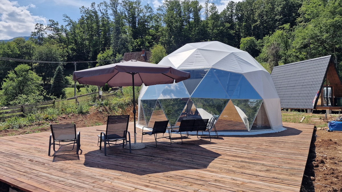 A geodesic dome structure features large transparent panels, allowing for ample natural light. Surrounding the dome is a spacious wooden deck equipped with several lounge chairs and a large umbrella, set against a backdrop of lush greenery and distant trees.