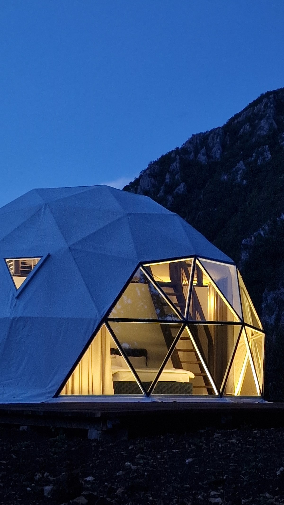 A geodesic dome is illuminated at dusk, showcasing large triangular windows that reflect the soft glow inside. A serene mountain backdrop underscores the unique architectural design, while the exterior presents a modern and minimalist aesthetic.