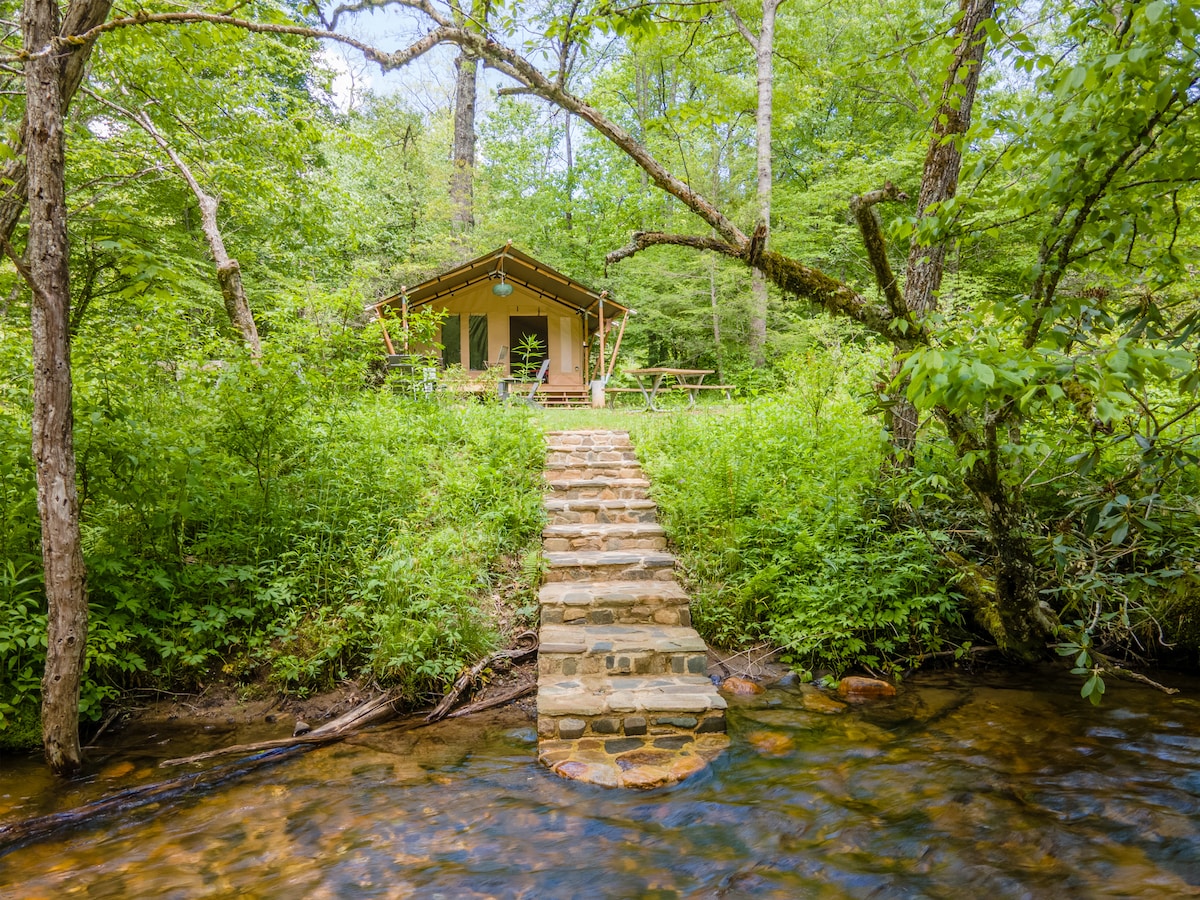 A natural stone pathway leads from the bank of the river to a canvas tent nestled amongst trees. The tent is surrounded by lush greenery, with picnic tables visible on the outdoor deck, inviting guests to enjoy the serene setting.
