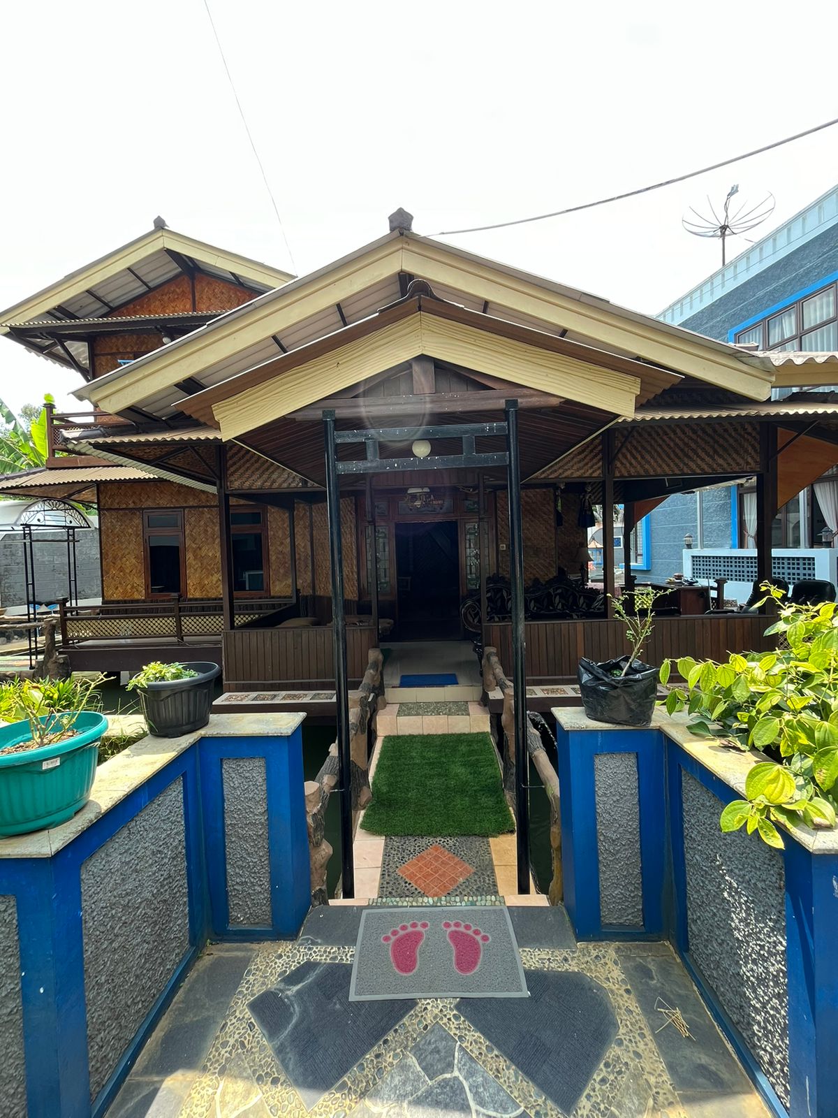 An inviting entrance is framed by decorative stone pathways and vibrant potted plants. A covered walkway leads to the open structure, characterized by traditional wooden architecture. Sunlight floods the space, creating a bright and welcoming ambiance.